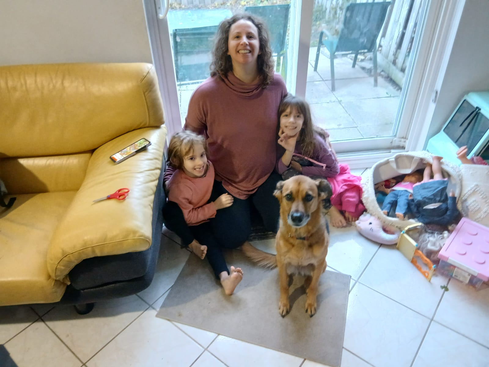 Aynav, co-owner of Dogs R The Best, sits on the floor at home with her two young daughters and their family dog, Roxy, all smiling together in a warm, natural family moment.